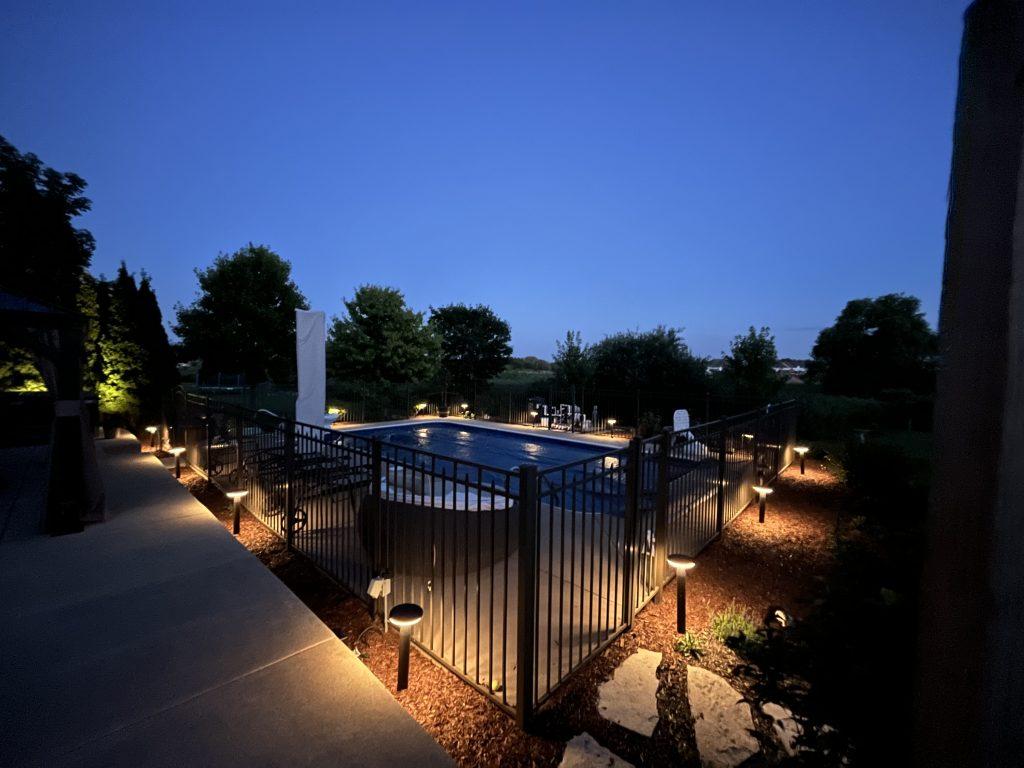 Outdoor lighting around a pool, patio, and walkway at dusk